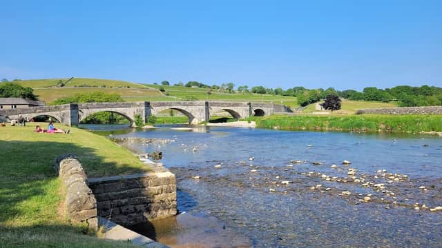 Scenic view of a stone bridge over a river, lush green hills, and a grassy area with people relaxing.