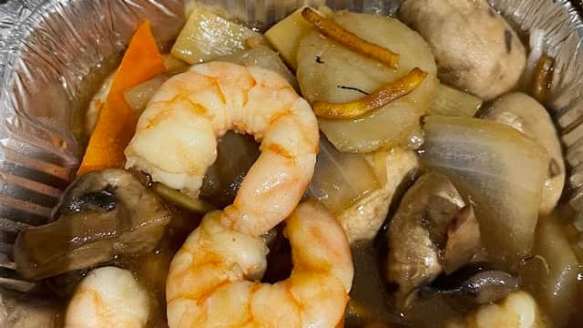 Shrimp and vegetables stir-fry in a foil container, featuring carrots, mushrooms, and onions in a savory sauce.