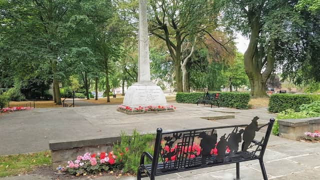 A memorial cross surrounded by trees and flower beds, with a decorative bench in the foreground.