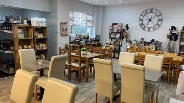 Cozy cafe interior with wooden tables, chairs, a large clock, and shelves displaying various products. Brightly lit space.