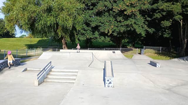 Skatepark with ramps, steps, and a smooth concrete surface, surrounded by trees and green grass.