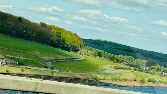Lush green hills and winding roads near Barkisland Park, with a scenic view of water and distant fields under a blue sky.