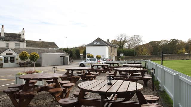 Outdoor seating area with wooden picnic tables, lanterns, and trees, set against a backdrop of a rustic building.