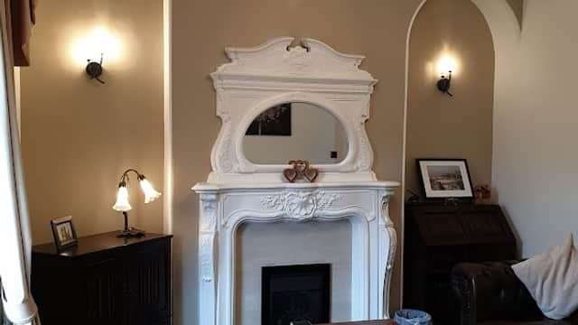 Cozy living room with a white fireplace, mirror, wooden table, leather sofa, and warm lighting.