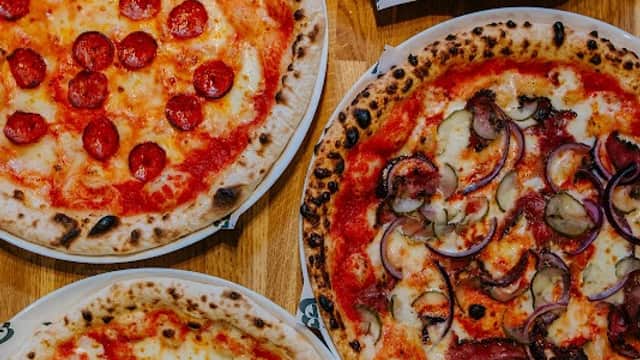 Four pizzas on wooden table: pepperoni, ham and mushroom, margherita with basil, and a cheesy pizza with red onions.