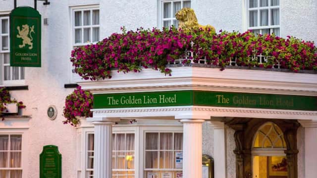 White hotel facade with floral balcony, green signage, golden lion statue, and arched entrance with columns.