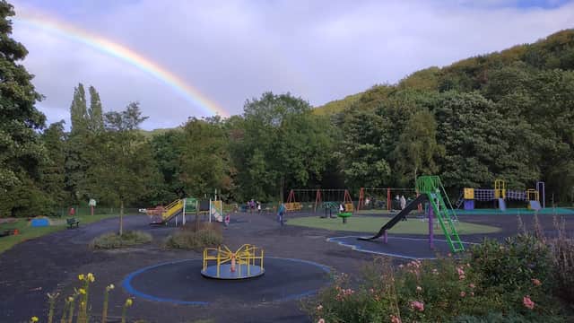 Colorful playground equipment under a rainbow, surrounded by green trees and blooming flowers in Graves Park, Woodseats.