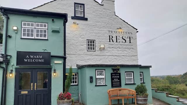 The Travellers Rest pub features a cozy exterior with a welcoming sign, flower pots, and a bench outside.