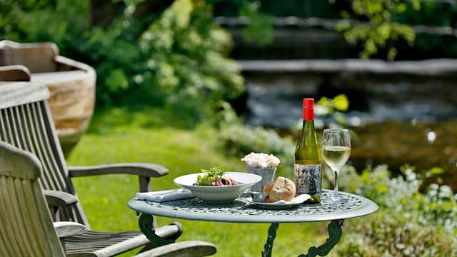 A table set with a salad, bread, a bottle of wine, and a glass, surrounded by greenery near a stream.