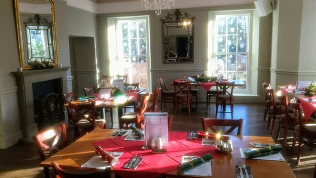 Bright dining area with wooden tables set with red napkins, a chandelier overhead, and large windows letting in natural light.