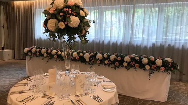 Elegant floral arrangements in tall vases and along a table, set in a softly lit room with sheer curtains.