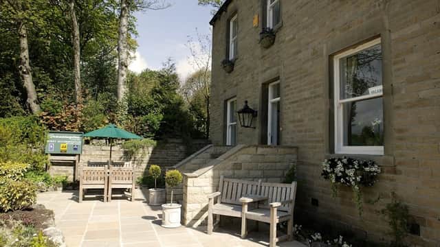 Charming stone building with a patio, benches, potted plants, and a shaded area in a lush, green setting.