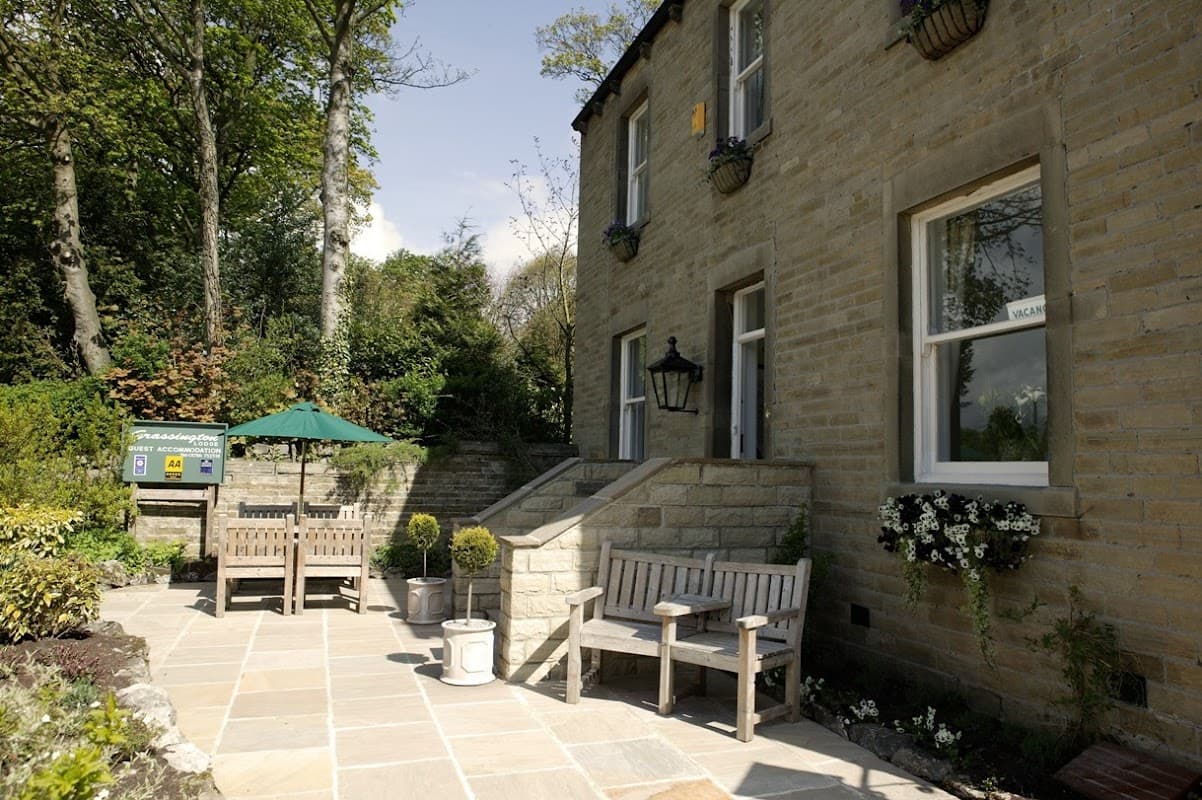 Charming stone building with a patio, benches, potted plants, and a shaded area in a lush, green setting.