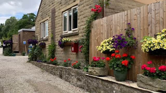 Charming stone building with vibrant flower pots and hanging baskets along a gravel pathway in a scenic campsite.
