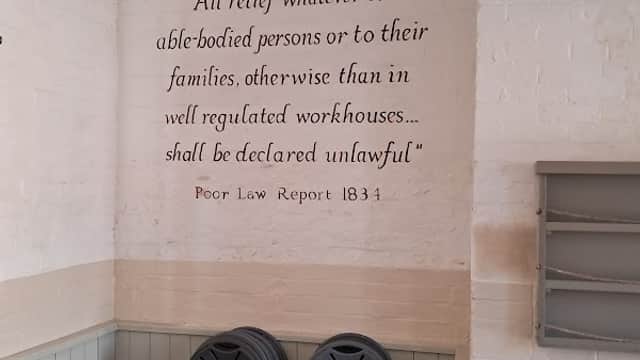 Text on wall quotes Poor Law Report 1834; stacked chairs and tables visible in a bright, airy car park space.