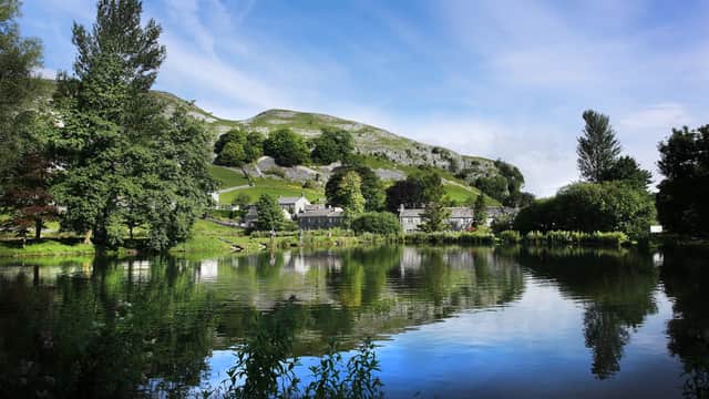 Tranquil lake reflecting lush trees and a hillside, with stone cottages nestled in the landscape under a blue sky.