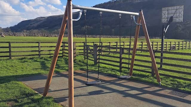 Wooden swingset in a grassy playground, surrounded by hills and a wooden fence under a blue sky with clouds.