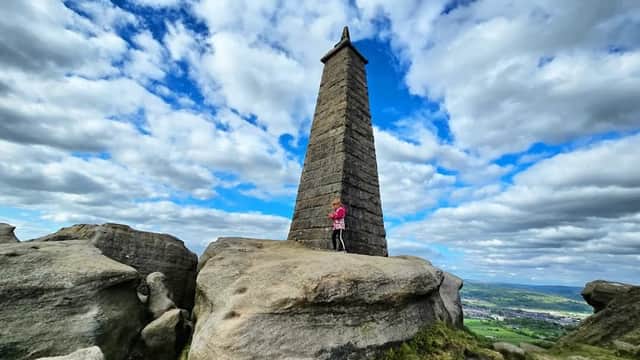 A tall stone monument stands on rocky terrain under a cloudy sky, with a person in pink near its base.
