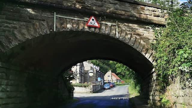 Stone bridge arching over a narrow road, with a warning sign and quaint buildings in the background under a blue sky.