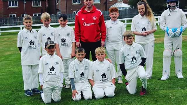 Youth cricket team in white uniforms poses on a grassy field, with a coach in a red jacket and a player in cricket gear.
