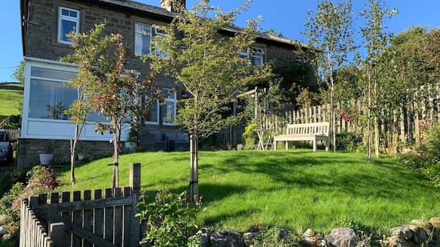 Fellside Cottage with a lush green garden, trees, and a wooden bench, set against a clear blue sky.
