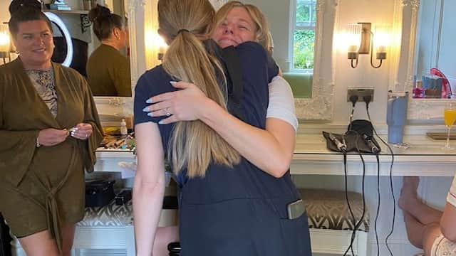 Two women embrace joyfully in a bright room with mirrors, while another woman watches nearby.