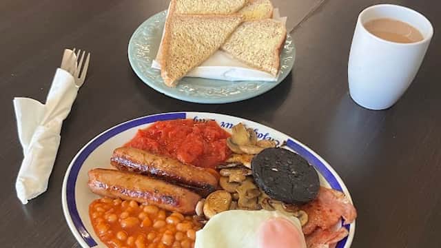 A full English breakfast with sausages, beans, mushrooms, bacon, a fried egg, toast, and a cup of tea on a table.