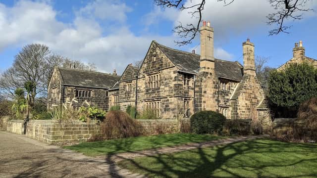 Historic stone building surrounded by lush greenery and trees under a partly cloudy sky. Pathway leads to the entrance.