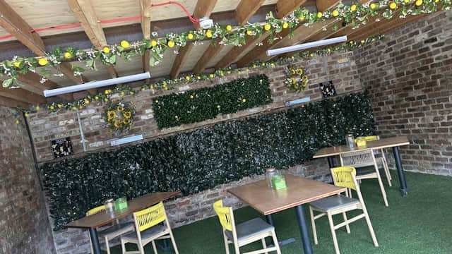 Rustic bar interior with wooden beams, greenery, and tables set on artificial grass. Brightly decorated with flowers.