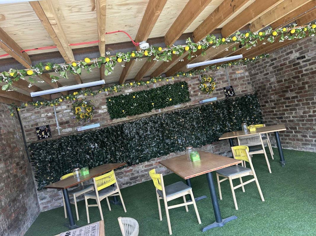 Rustic bar interior with wooden beams, greenery, and tables set on artificial grass. Brightly decorated with flowers.