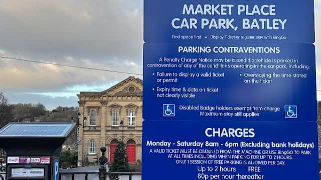 Sign for Market Place Car Park in Batley, displaying parking rules, charges, and payment options. Historic building in background.