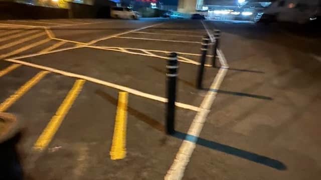 Dark car park with yellow lines, black bollards, and illuminated buildings in the background. Nighttime setting.