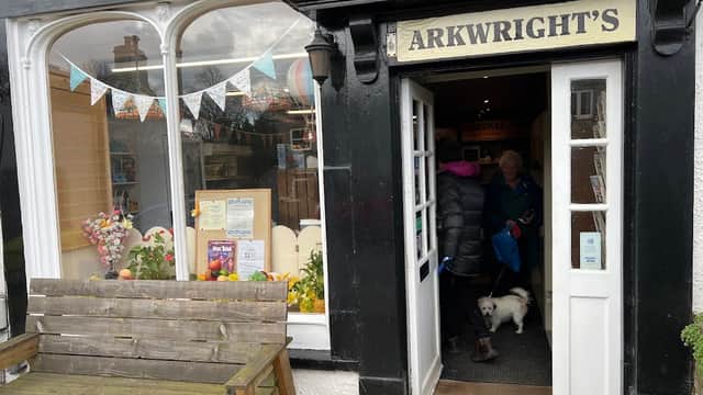 Quaint village shop entrance with a wooden bench, colorful bunting, and customers entering with a small dog.