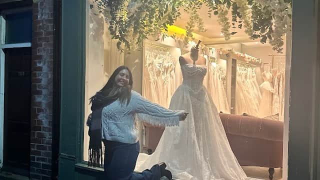 A woman poses joyfully outside a bridal shop featuring elegant wedding dresses in the window display.