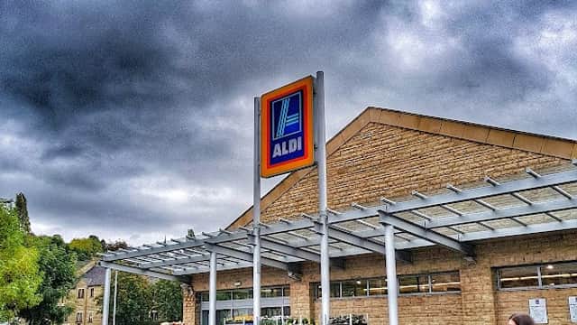 ALDI storefront in Batley, Yorkshire, with shoppers and a cloudy sky overhead.