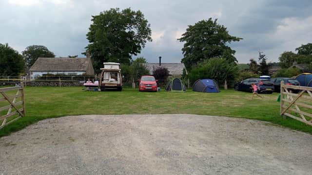 A grassy area with tents, parked cars, and a caravan, surrounded by trees and a stone building under a cloudy sky.