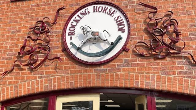 Sign for "The Rocking Horse Shop" on a brick building, featuring a rocking horse design and decorative swirls.