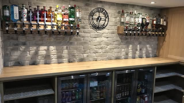 Bar area with a wooden counter, brick wall, and shelves displaying various bottles of spirits and a clock.
