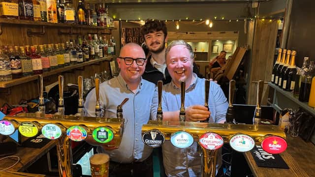 Two smiling men stand behind a bar with various beer taps and bottles, while a third man smiles in the background.