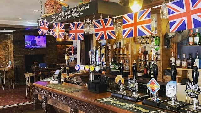 Wooden bar with British flags, shelves of spirits, and a TV screen in a cozy pub setting.