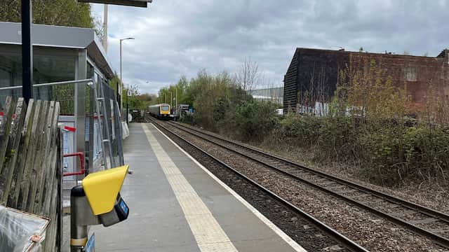 Platform with a "Way out" sign, rail tracks, greenery, and a train approaching in the distance under a cloudy sky.