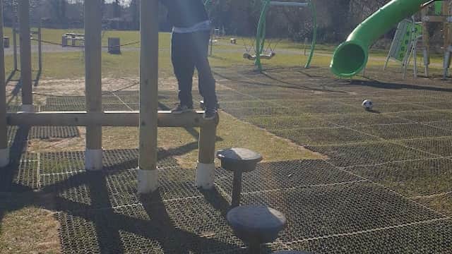 A child climbing on wooden playground equipment with a slide in the background, set in a grassy area under clear blue skies.