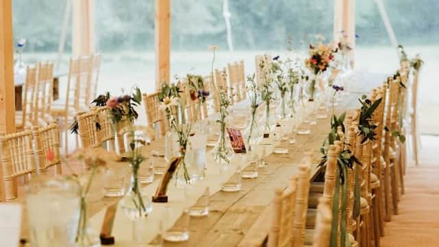 Long wooden table set with vases of flowers under a tent, surrounded by chairs and soft lighting.