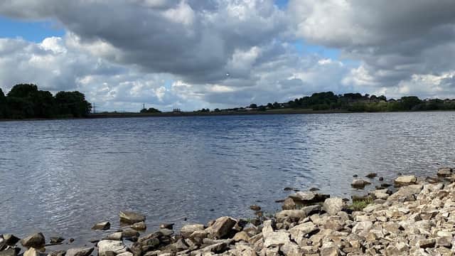 Calm water of Ardsley Reservoir surrounded by rocky shoreline and lush greenery under a cloudy blue sky.