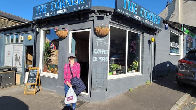 A woman stands outside "The Corner," a Greek and Turkish restaurant with a blue sign and potted plants.