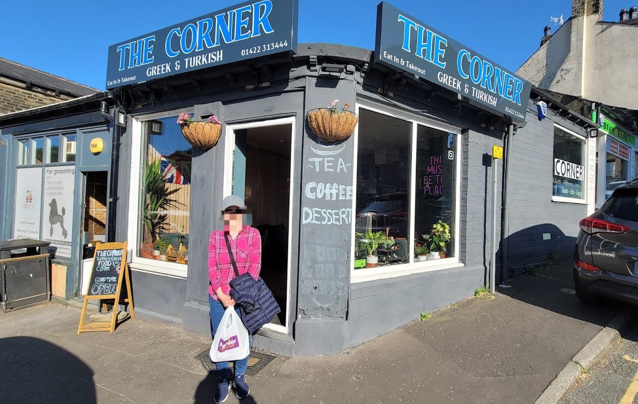 A woman stands outside "The Corner," a Greek and Turkish restaurant with a blue sign and potted plants.