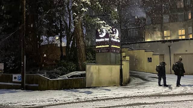 Snow-covered ground with two people walking, streetlights illuminating a multi-storey car park entrance in Harrogate.