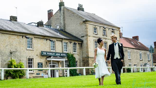 Couple walking hand-in-hand on green lawn in front of the Worsley Arms Hotel, a charming stone building.