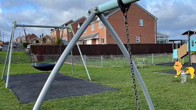 Playground with swings, a spring rider, and grassy areas, surrounded by houses under a blue sky with clouds.
