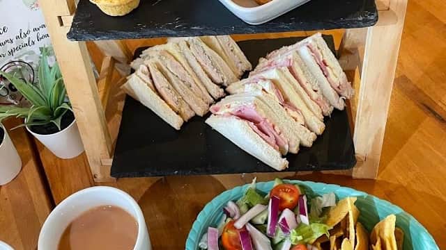 A wooden display holds sandwiches, pastries, and a bowl of sausages, with a side of salad and a cup of tea on the table.